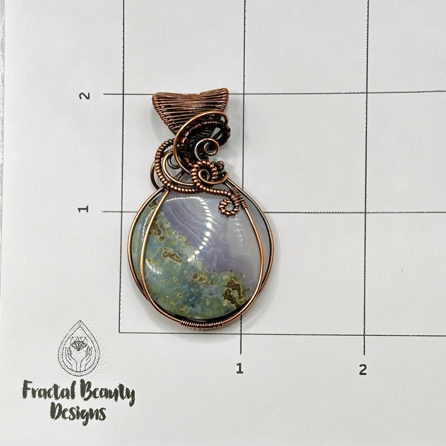 Circular Indonesian Moss Agate stone wrapped in antiqued copper on measurement grid