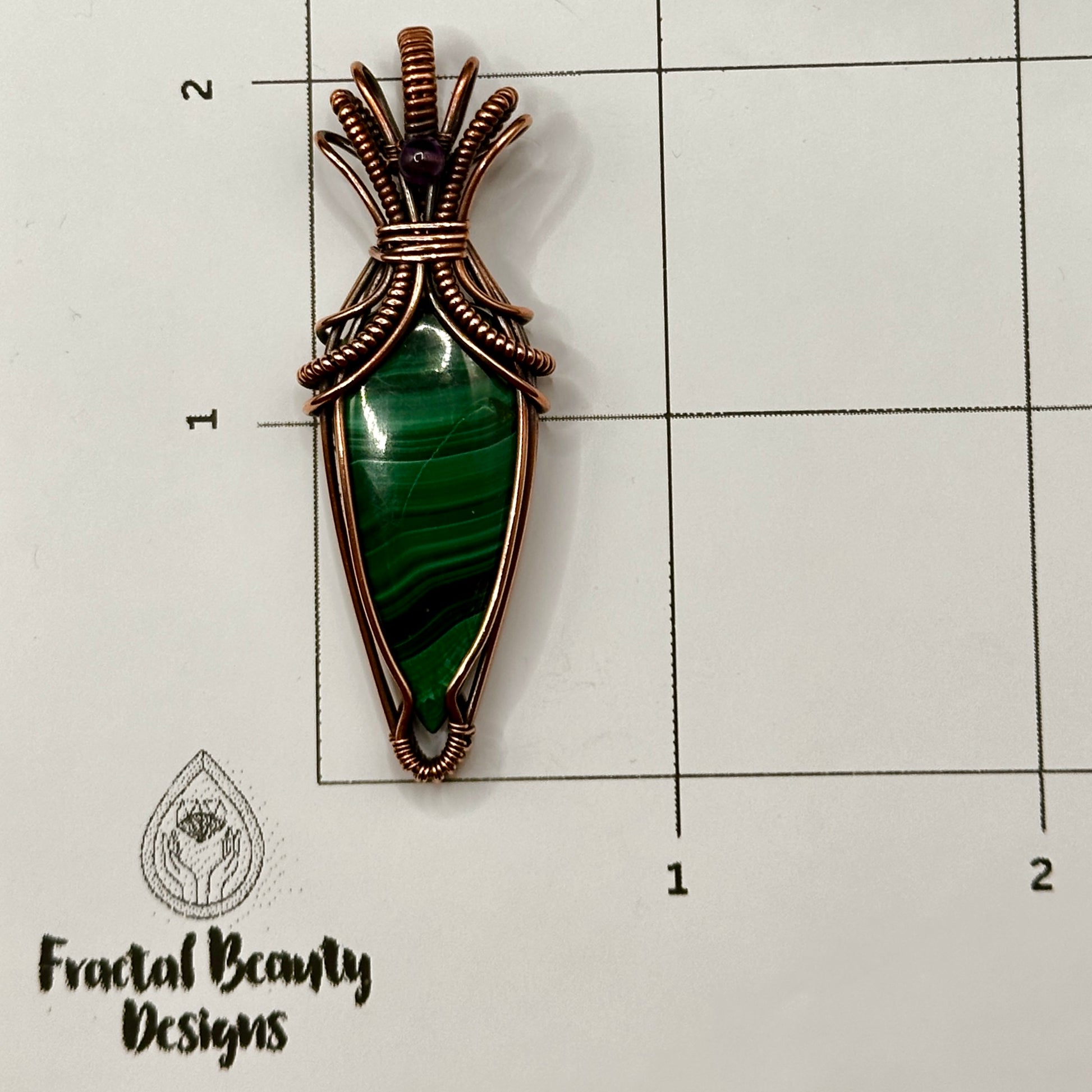 Tear shaped malachite stone wrapped in an "art deco" design with copper wire and accented with an amethyst bead on measuring grid